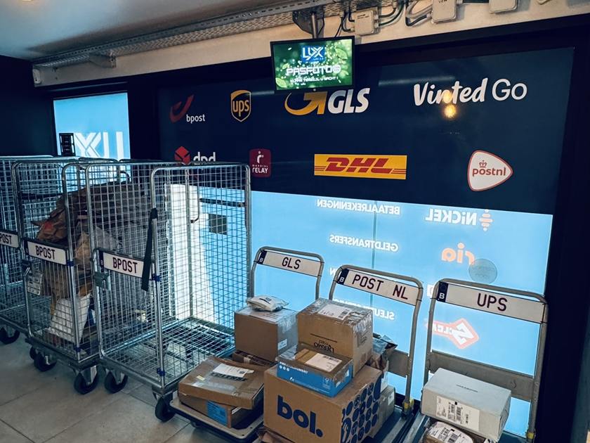 Het pakjesdepot van LUX 2.0 met de logowand van alle acht koeriers en de rolcontainers vol pakketten